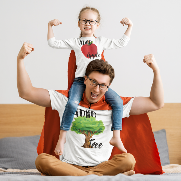 A photo of a man with his daughter on his shoulders, both of them flexing their arms wearing red capes and white t-shirts. The white t-shirts display some of Julia’s merchandise designs . The dads t-shirt has a tree on it with the text “ADHD” above the tree and “tree” below the tree. The daughters t-shirt has an apple on it with the text “ADHD” above the apple and “apple” below the apple making a play on the phrase the apple doesn't fall far from the tree, presenting the dad as the ADHD tree and the daughter as the ADHD apple.