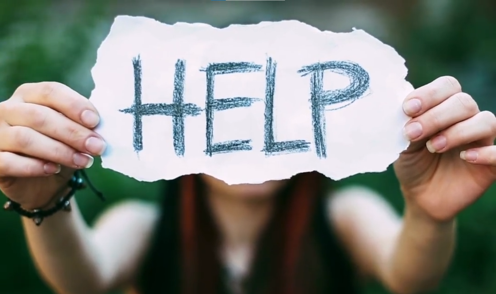 A Young adult holds up a piece of paper with the word help hand written on it. The piece of paper obscures their face. They have long reddich colour hair.