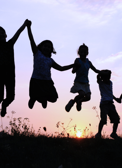 Children holding hands and helping each other jump high. They are sillhouetted against a sunset.